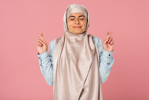 boldog Arab lány hidzsáb ujjal keresztbe, és csukott szemmel, izolált rózsaszín  - Fotó, kép