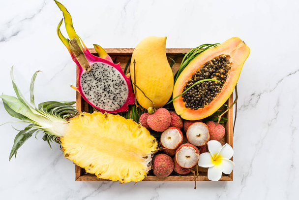 Fresh exotic fruits in wooden box on white marble background - sliced papaya, mango, pineapple, dragon fruit, lychee. Mockup, flat lay, overhead. Top view. - Foto, imagen