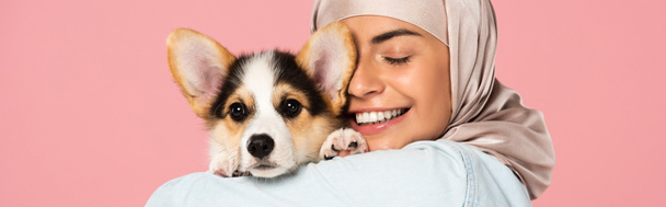 smiling muslim woman in hijab holding Welsh Corgi puppy, isolated on pink - Fotoğraf, Görsel
