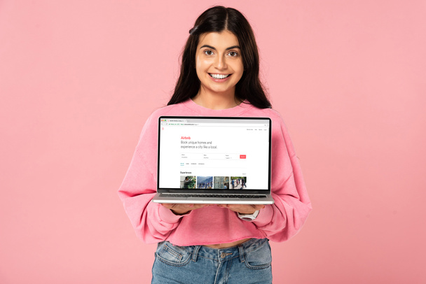 KYIV, UCRANIA - 30 de julio de 2019: niña sonriente sosteniendo el ordenador portátil con airbnb sitio web en la pantalla, aislado en rosa
 - Foto, Imagen