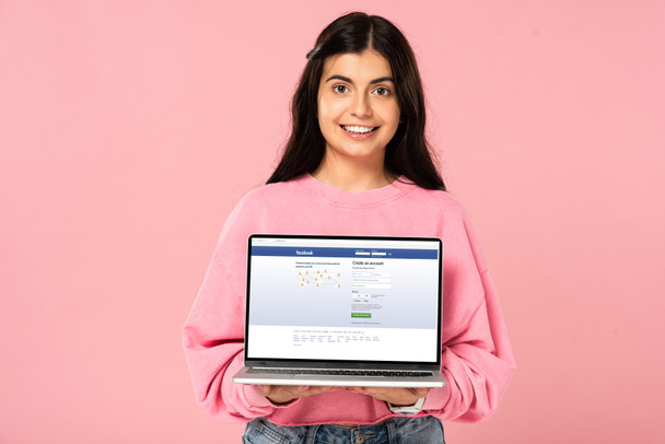 KYIV, UCRANIA - 30 de julio de 2019: niña sonriente sosteniendo el ordenador portátil con el sitio web de facebook en la pantalla, aislado en rosa
 - Foto, Imagen