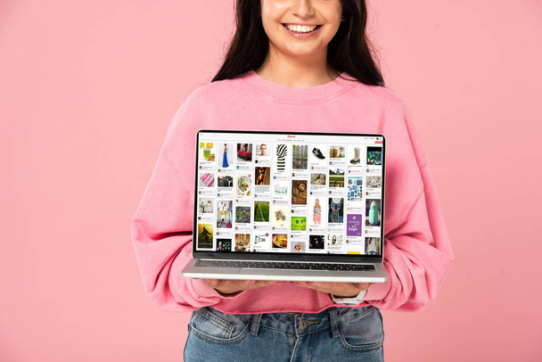 KYIV, UKRAINE - JULY 30, 2019: cropped view of smiling girl holding laptop with pinterest website on screen, isolated on pink - 写真・画像