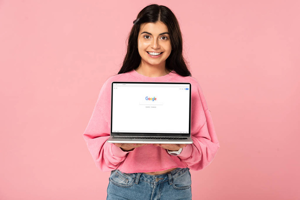 KYIV, UKRAINE - JULY 30, 2019: smiling girl holding laptop with google website on screen, isolated on pink - Фото, изображение