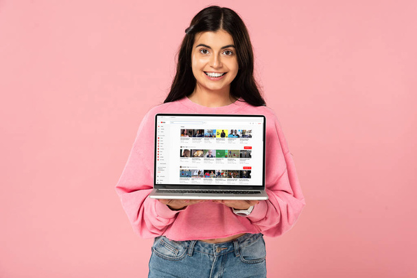 KYIV, UKRAINE - JULY 30, 2019: smiling girl holding laptop with youtube website on screen, isolated on pink - Foto, Imagem