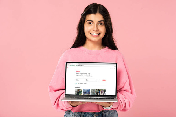KYIV, UKRAINE - JULY 30, 2019: smiling girl holding laptop with airbnb website on screen, isolated on pink - Zdjęcie, obraz
