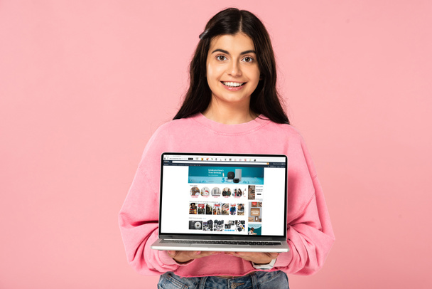 KYIV, UKRAINE - JULY 30, 2019: smiling girl holding laptop with amazon website on screen, isolated on pink - Фото, изображение