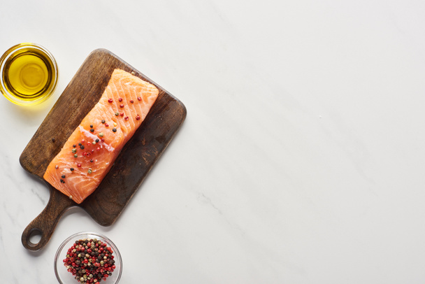 top view of raw salmon steak with peppercorns on wooden cutting board near oil on marble surface - Fotoğraf, Görsel