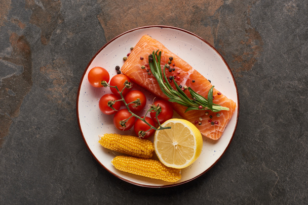 top view of raw salmon steak with tomatoes, corn, lemon, rosemary and pepper on plate - Foto, afbeelding