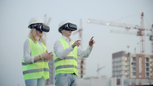 zwei Personen in Virtual-Reality-Brillen auf dem Hintergrund von im Bau befindlichen Gebäuden mit Kränen imitieren die Arbeit der Schnittstelle zur Steuerung und Verwaltung von Bauvorhaben - Filmmaterial, Video
