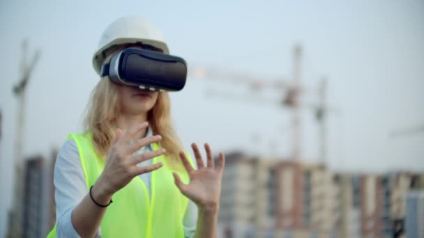 Жінка-дизайнер на будівельному майданчику в твердій шапці і жилеті в окулярах віртуальної реальності, щоб перемістити руки імітуючи інтерфейс на тлі кранів на заході сонця
 - Кадри, відео