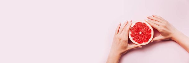 Female's hands holding grapefruit on pink background with tropic plants and leaves. - Foto, Bild