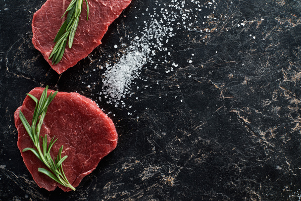 top view of raw beef steaks with rosemary twigs on black marble surface with scattered salt crystals - Φωτογραφία, εικόνα