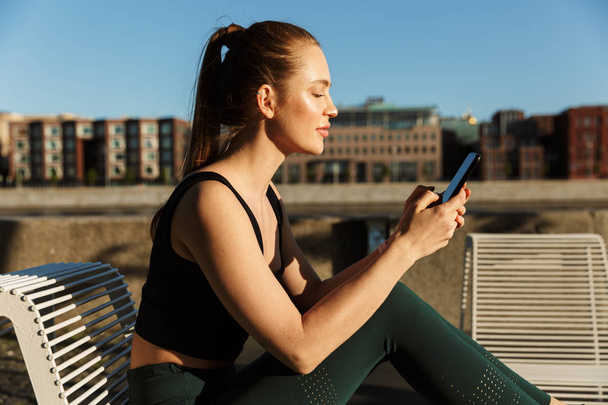 Image of sunlit sporty woman holding smartphone and sitting on c - Foto, imagen