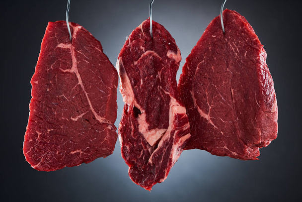 rohe Rindfleischstücke auf Metallhaken auf dunkelschwarzem Hintergrund - Foto, Bild