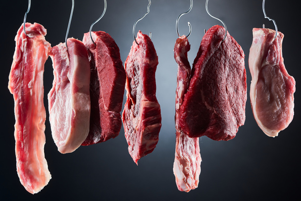 assorted raw meat and bacon on metal hooks on dark black background - Фото, изображение