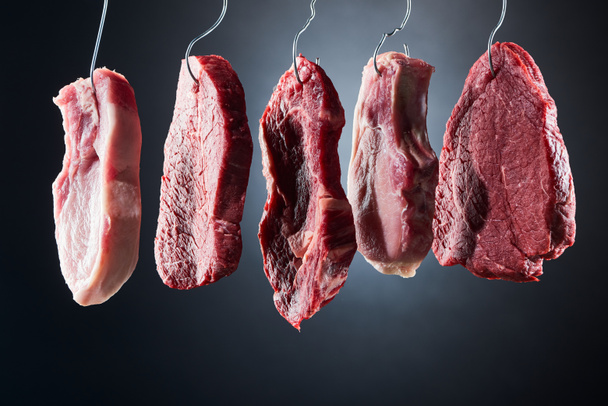 різноманітне сире м'ясо на металевих гачках на темно-чорному тлі
 - Фото, зображення