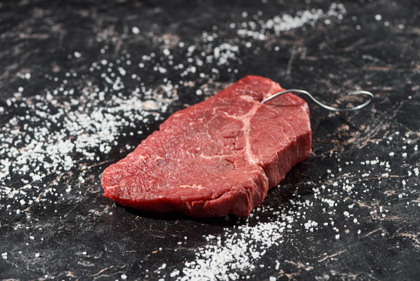 raw meat steak with metal hook on black marble surface with scattered salt  - Fotografie, Obrázek