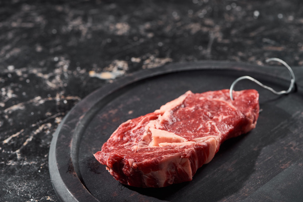 raw meat with hook on wooden plate on black marble surface - Фото, изображение