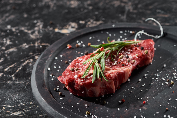 raw meat steak with rosemary twig sprinkled with salt and pepper on round wooden surface - Photo, Image