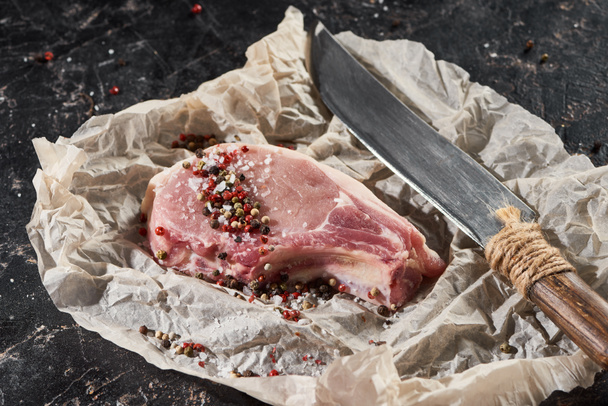 raw pork steak sprinkled with salt and pepper near knife on parchment paper on black marble surface - Foto, Imagem