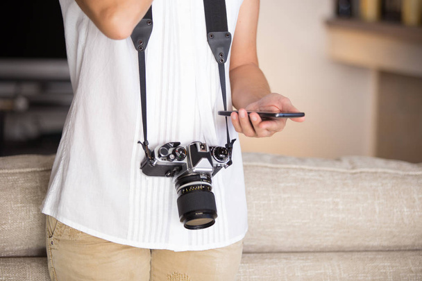 Contraste entre os tempos antigos e modernos: uma jovem com uma câmera vintage ao redor do pescoço mexe com seu smartphone
 - Foto, Imagem