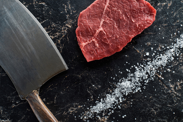 ナイフの近くに調理されていない牛肉サーロインのトップビューと黒い大理石の背景に散らばった塩 - 写真・画像