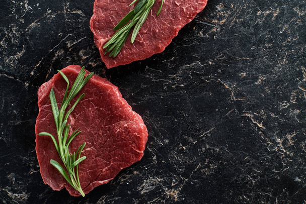 top view of uncooked beef sirloins with rosemary on black marble background - Fotoğraf, Görsel