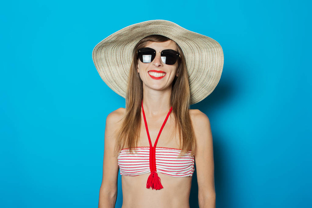 Young woman in a swimsuit, glasses and a wide-brimmed hat on a blue background. Concept of vacation, summer, vacation at sea - Photo, Image