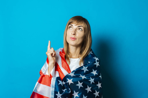 junge Frau in US-Flagge gehüllt mit nachdenklichem Gesicht auf blauem Hintergrund. Visa-Konzept, gute Idee, Fragezeichen, Frage - Foto, Bild