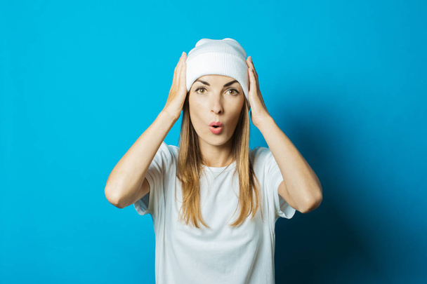 Mujer joven con un sombrero blanco y una camiseta con una cara sorprendida sobre un fondo azul
 - Foto, Imagen