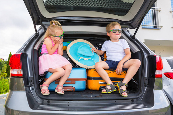 Gemelos niño y niña están sentados en el maletero del coche con el equipaje, listo para ir de vacaciones
. - Foto, Imagen