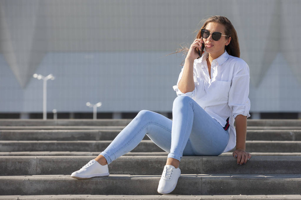Young American woman in a white T-shirt in jeans in white trendy sneakers relaxes sitting on the steps. Cute european girl model enjoys the rest. Spring style women's clothing. - Zdjęcie, obraz