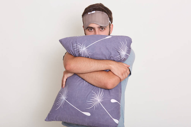 Studio shot of young handsome guy on white background wearing t shirt, holding pillow in his hands, male hugging his pillow, ready for falling asleep, man posing with blindfold on his forehead. - Foto, Bild