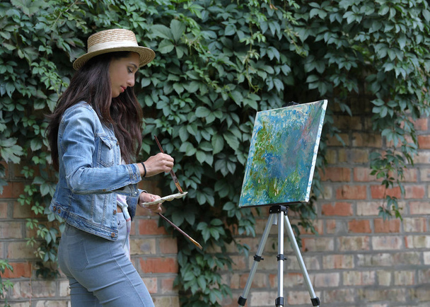 Jovem bela mulher de 30 anos em um chapéu pintando um quadro usando escovas e um cavalete moderno na natureza em um parque de verão perto de uma parede de tijolo
 - Foto, Imagem