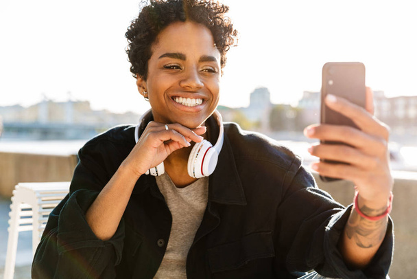 Portrait of cheerful african american woman with headphones making video call on smartphone near riverfront - Fotoğraf, Görsel