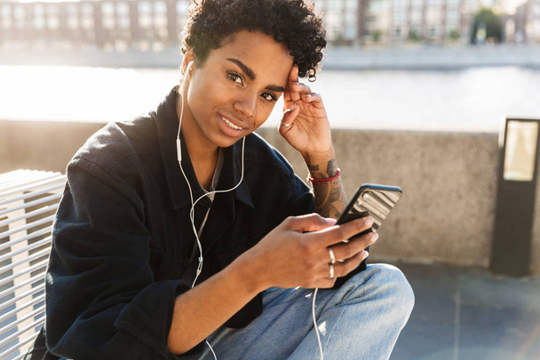 Portrait closeup of beautiful african american woman listening to music on cellphone with earphones near riverfront - Foto, imagen