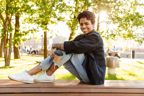 Photo of cheerful african american woman holding paper cup while sitting on bench in city park - Valokuva, kuva