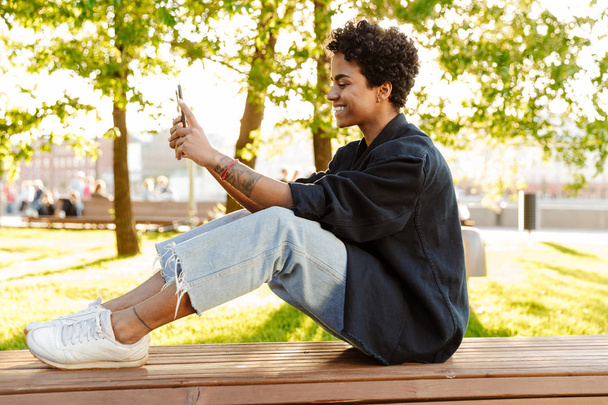 Photo of pleased african american woman taking selfie photo on smartphone while sitting on bench in city park - Fotoğraf, Görsel