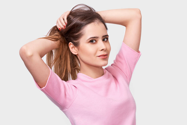 Portrait of attractive young woman smiling, wearing pink t-shirt, posing against white background. Pretty female has joyful expression, making ponytail hairstyle, poses in studio. - Photo, Image