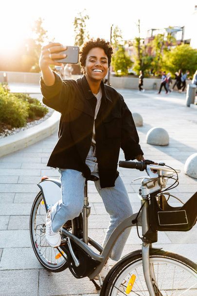 Photo of adorable african american woman taking selfie on cellphone while sitting on bicycle in city park - Fotografie, Obrázek