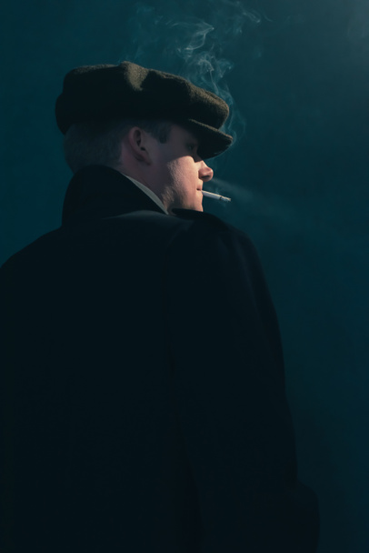 Ρετρό άντρας με καπέλο καπνίζει τσιγάρο σε δωμάτιο καπνού. Πλευρική όψη. - Φωτογραφία, εικόνα