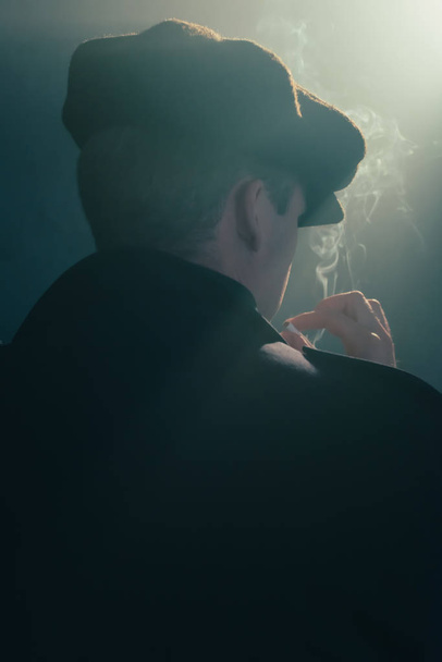 Hombre retro con gorra fuma cigarrillo en habitación ahumada. Vista trasera
. - Foto, Imagen