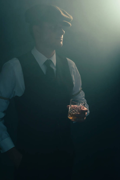 Retro businessman with cap holds glass of whiskey in smoky room. - Фото, зображення