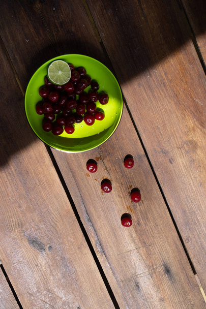 Green bowl in the upper left corner. It has cherries and lime. Several berries are scattered on a dark wood surface. Vertical orientation - Foto, Bild