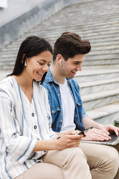 Happy young amazing loving couple business people colleagues outdoors outside on steps using mobile phone and laptop computer listening music with earphones. - Φωτογραφία, εικόνα