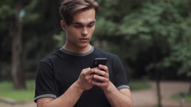 Handsome Young Man Looking Amazed Scrolling the Phone and Saying Wow Standing in Park. - Séquence, vidéo