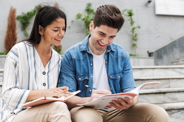 Image of smiling couple talking and studying while sitting on bench near stairs outdoors - Foto, imagen