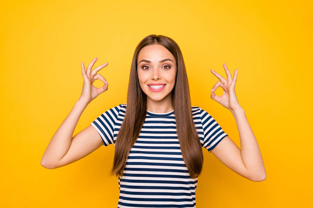 Portrait of cute pretty millennial in striped t-shirt showing ok sign advertising isolated over yellow background - Foto, imagen