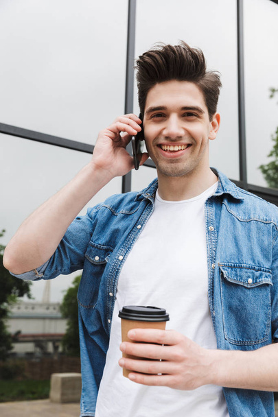 Щасливий молодий дивовижний чоловік бізнесмен позує на відкритому повітрі, ходячи по мобільному телефону, п'є каву
. - Фото, зображення