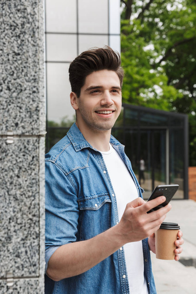 Onnellinen nuori hämmästyttävä mies liikemies poseeraa ulkona ulkona kävely chattailuun matkapuhelimella kahvin juominen
. - Valokuva, kuva